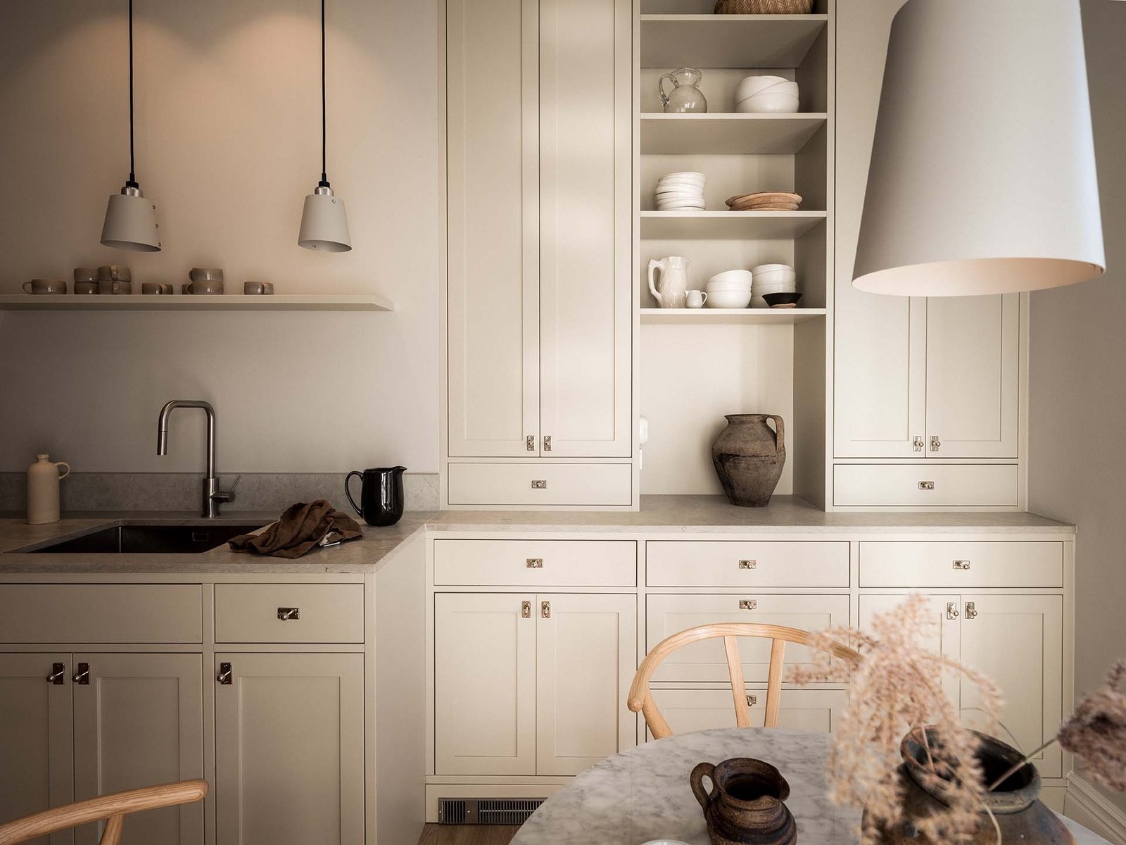 Öppna hyllor med porslin, vattenkokare vid handfat och runt bord med torkade blommor under Buster and Punch-pendlar